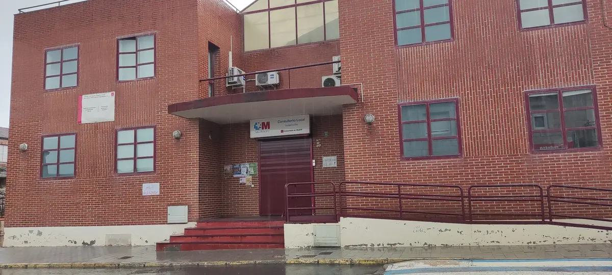 The brick entrance of a Consultorio Local, a village public health center in the Madrid region, with a ramp and red steps on a rainy morning.